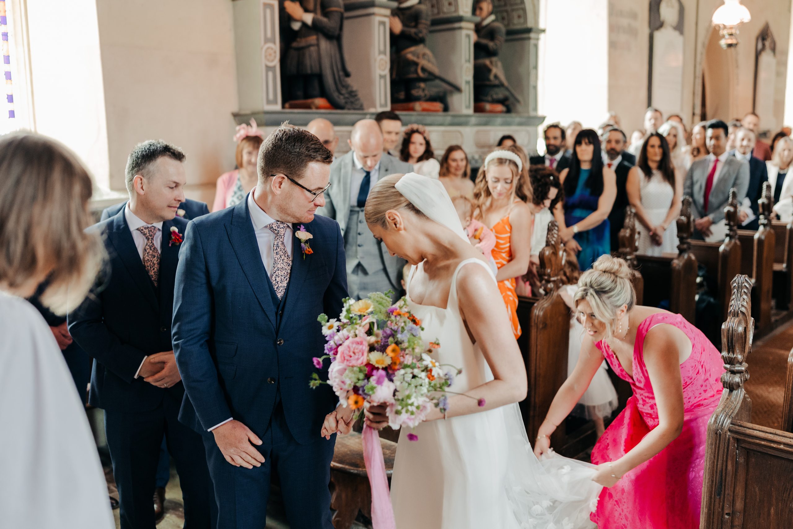 Bride and groom stood at the front of the aisle with maid of honour adjusting the bride's Suzanne Neville wedding gown, coordinated by Hardesty Event Design