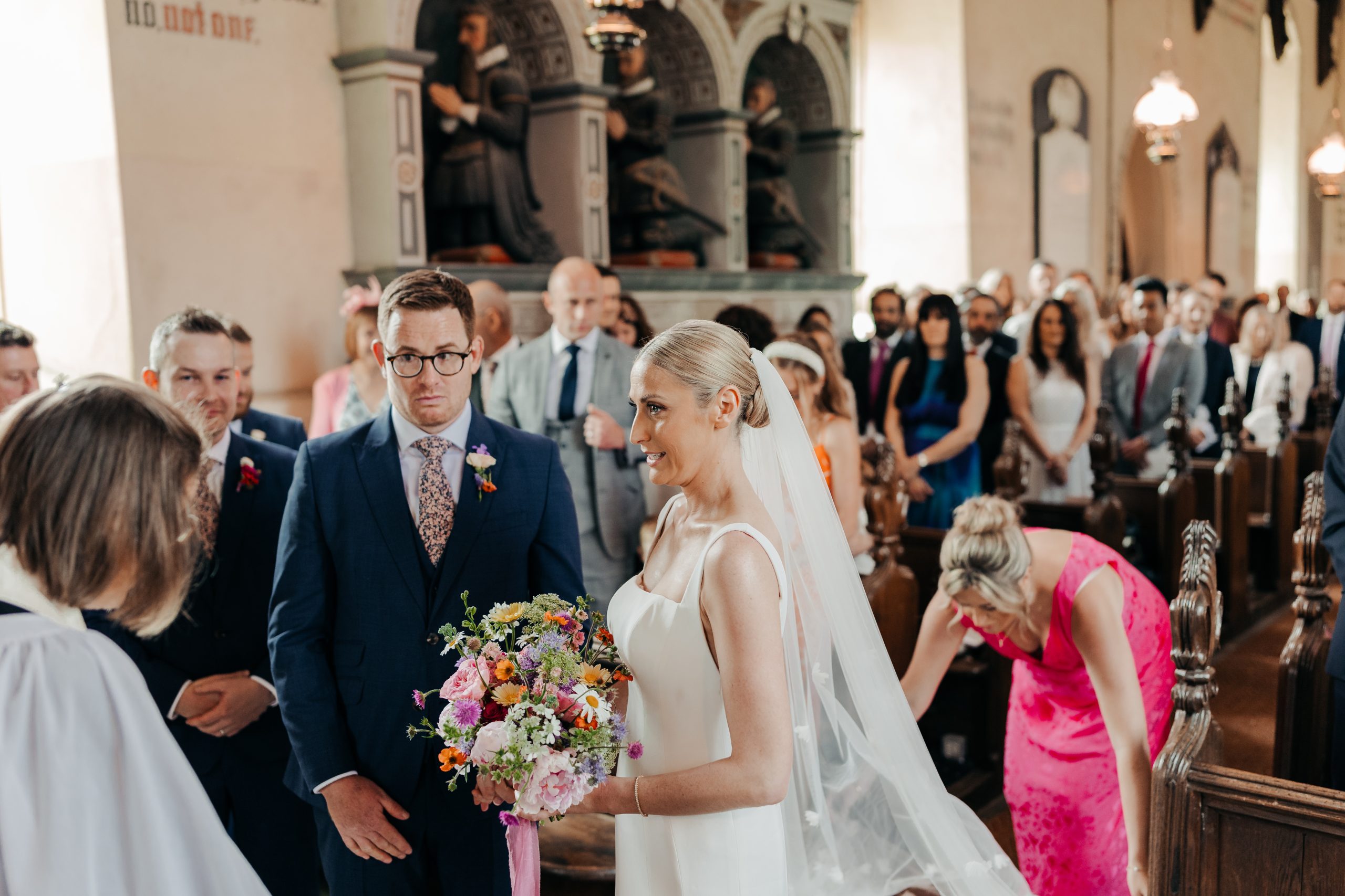 Bride and groom stood at the front of the aisle with maid of honour adjusting the bride's Suzanne Neville wedding gown, coordinated by Hardesty Event Design
