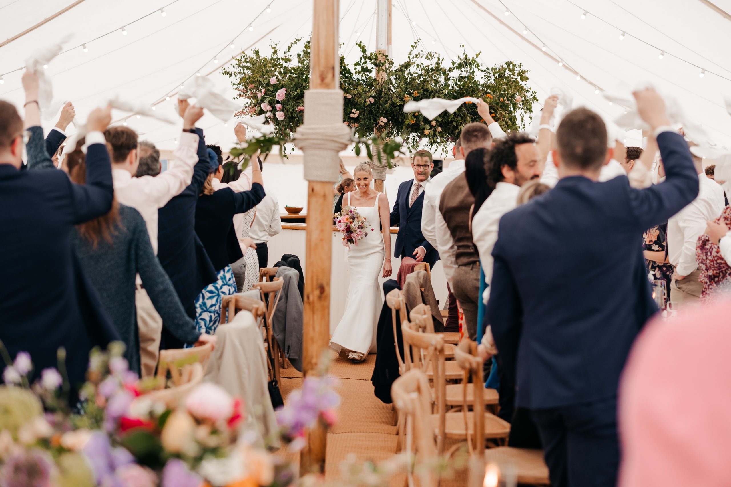 bride and groom welcomed into their marquee wedding reception by guests and beautiful floral arrangements suspended above, planned by professional wedding planners Hardesty Event Design