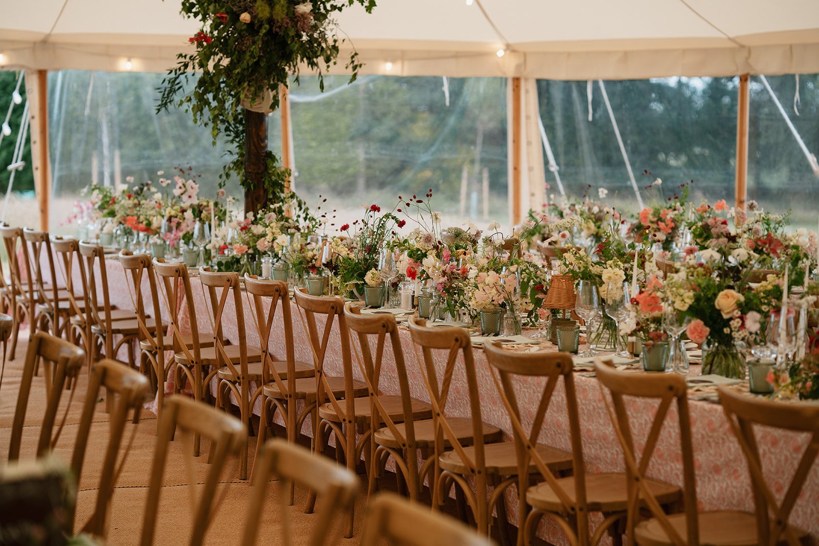 Long tables with patterned tablecloths under marquee with hand-printed textiles, candles and coral, cream and red floral centrepieces at a wedding in Lincolnshire