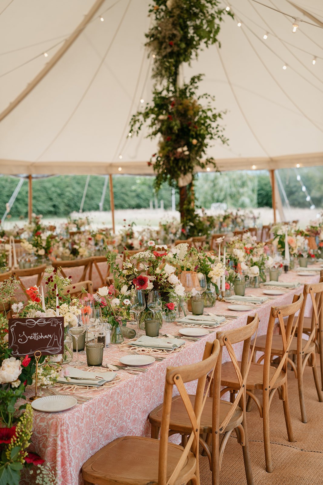 Soft abundant flowers in shades of cream, coral and red, arranged along the length of a marquee table and up the poles of the marquee. Styled by UK wedding planner