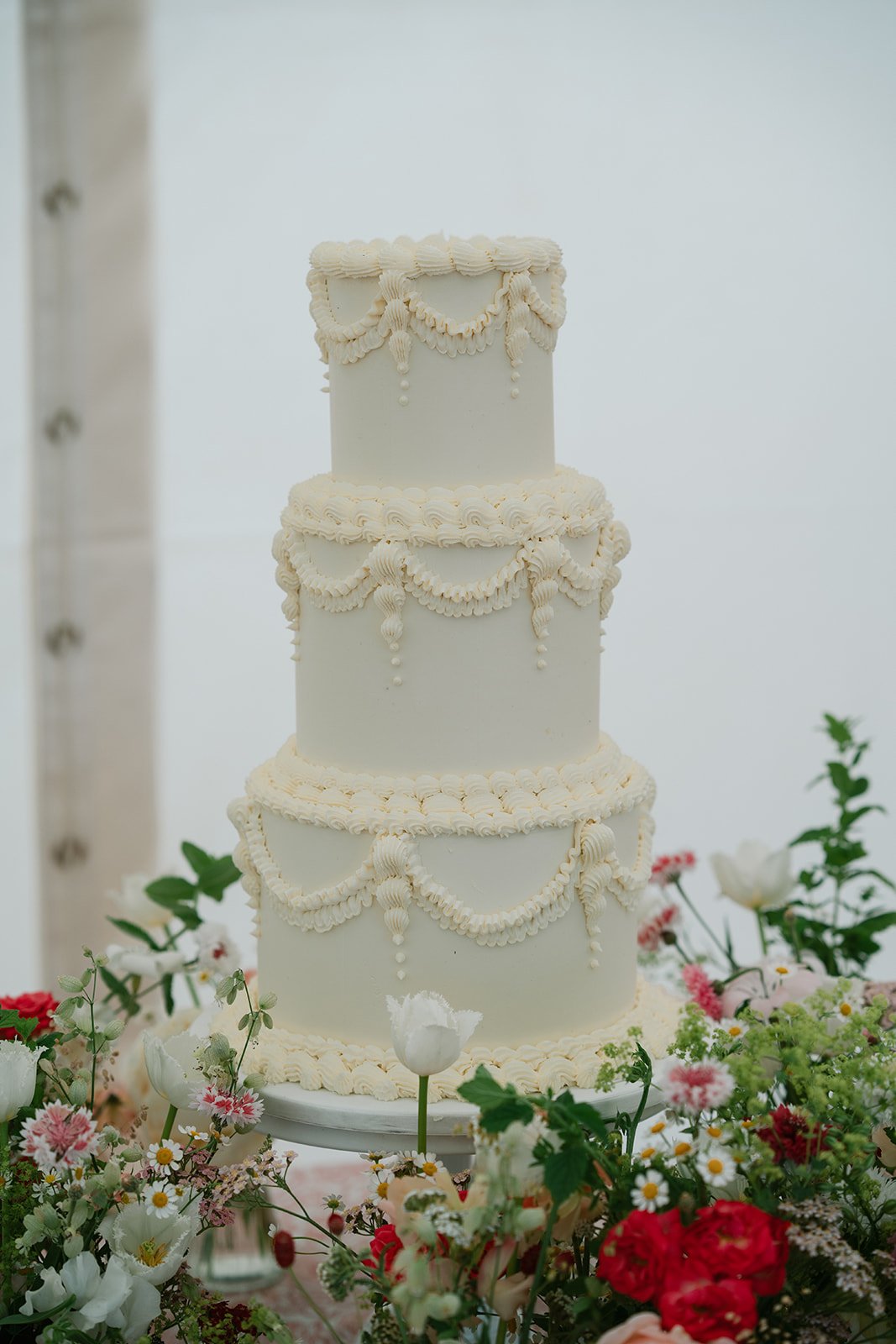 white iced wedding cake with a bed of flowers to complement sat below on charlotte and ed's wedding day, styled by UK wedding planners Hardesty Event Design